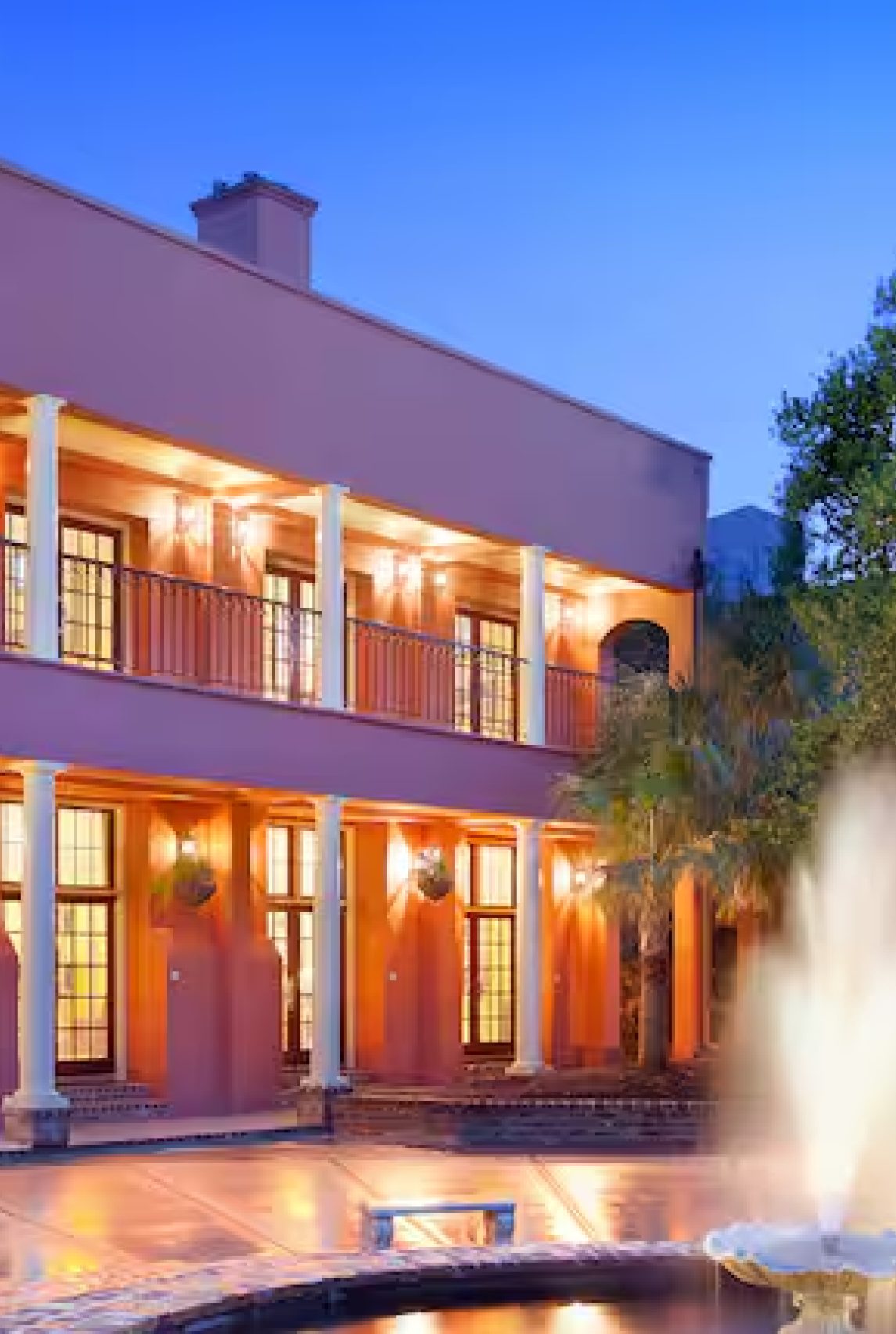 A grand, multi-story pink building with illuminated balconies and a vibrant fountain in foreground at twilight.