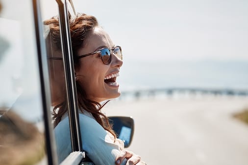 Joyful woman in sunglasses laughing and leaning out of a car window on a sunny road trip.