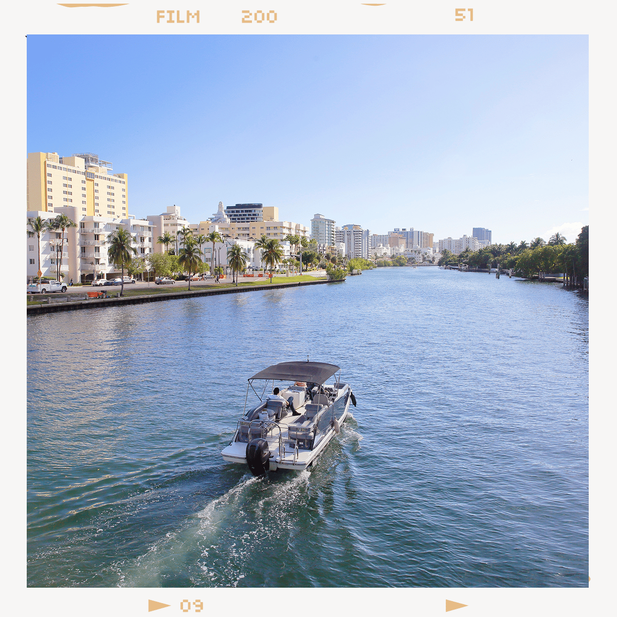 A pontoon boat motors down a bright blue canal lined with palm trees and a city skyline under a clear sky.