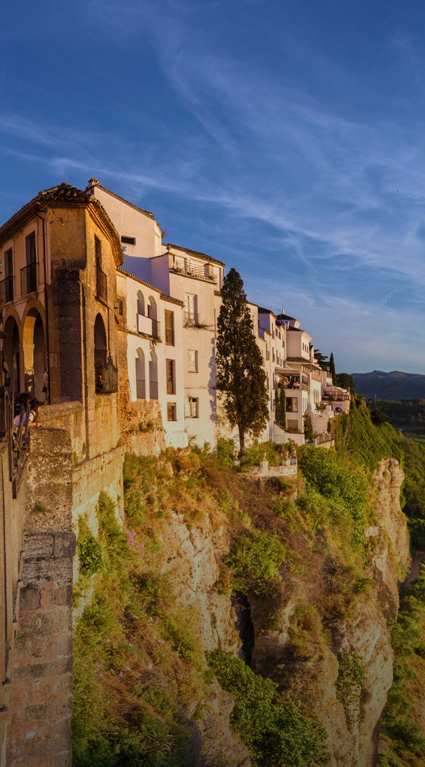 White buildings perched on a dramatic cliffside overlooking a valley under a blue sky.