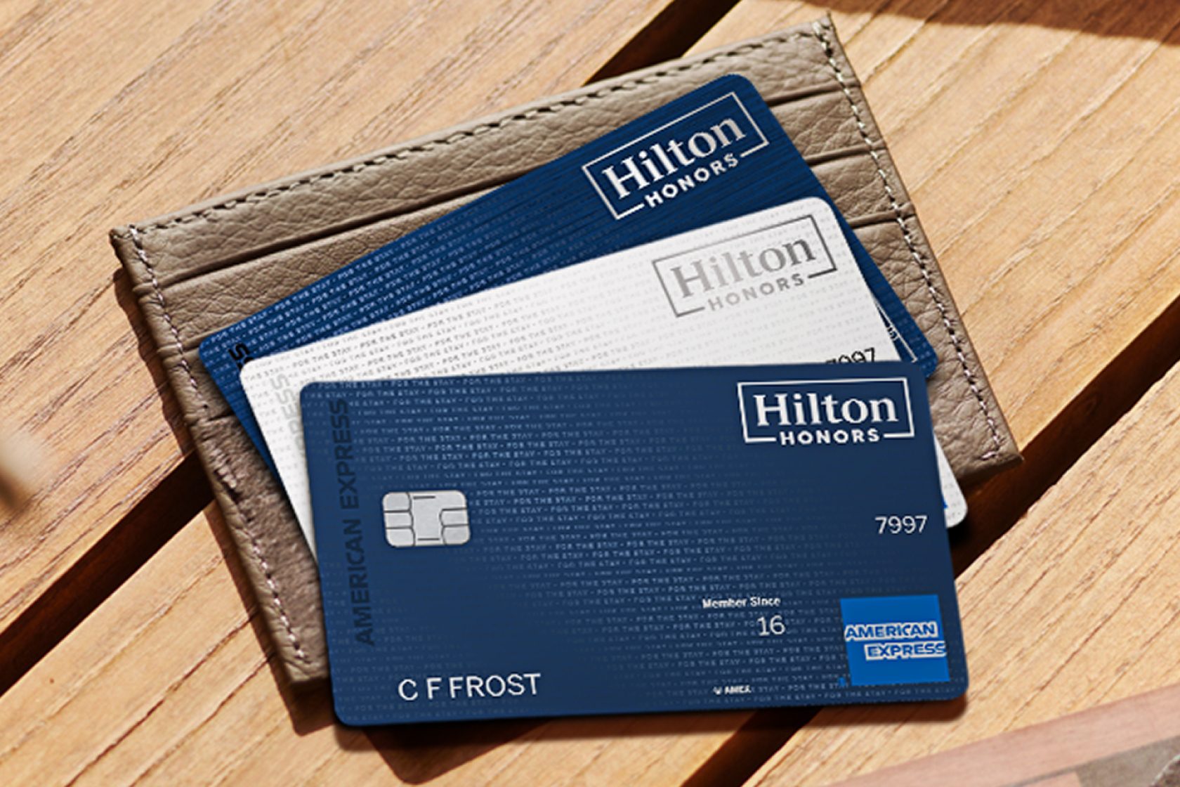 Three Hilton Honors American Express cards, blue and white, stacked in a leather card holder on a wooden table.