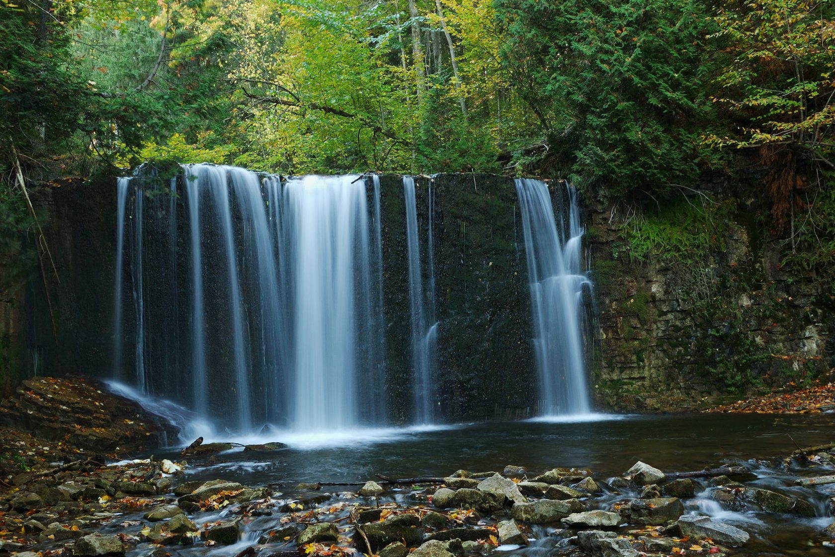 A long-exposure photograph of a waterfall cascading into a rocky stream, surrounded by lush green trees.