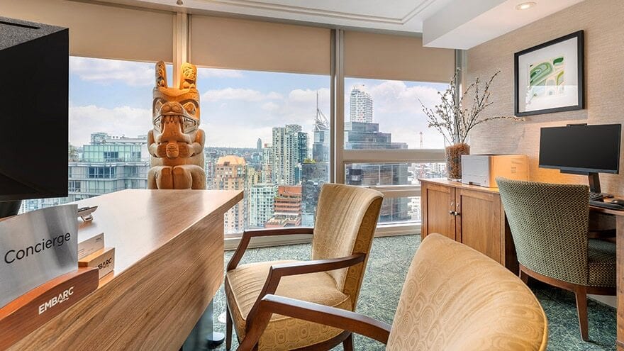 An office space with a concierge desk, a wooden totem pole, two chairs, and a computer, overlooking a city.