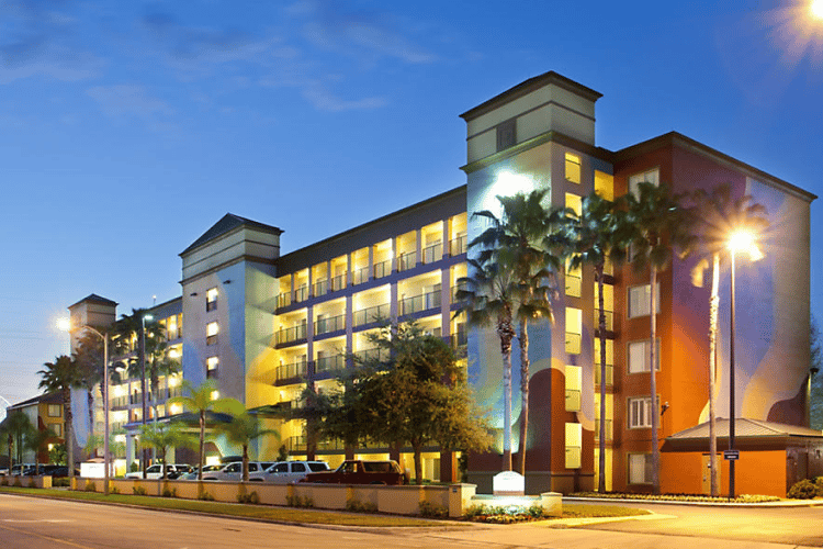 Colorful resort hotel illuminated at dusk with palm trees and parked cars along a street.