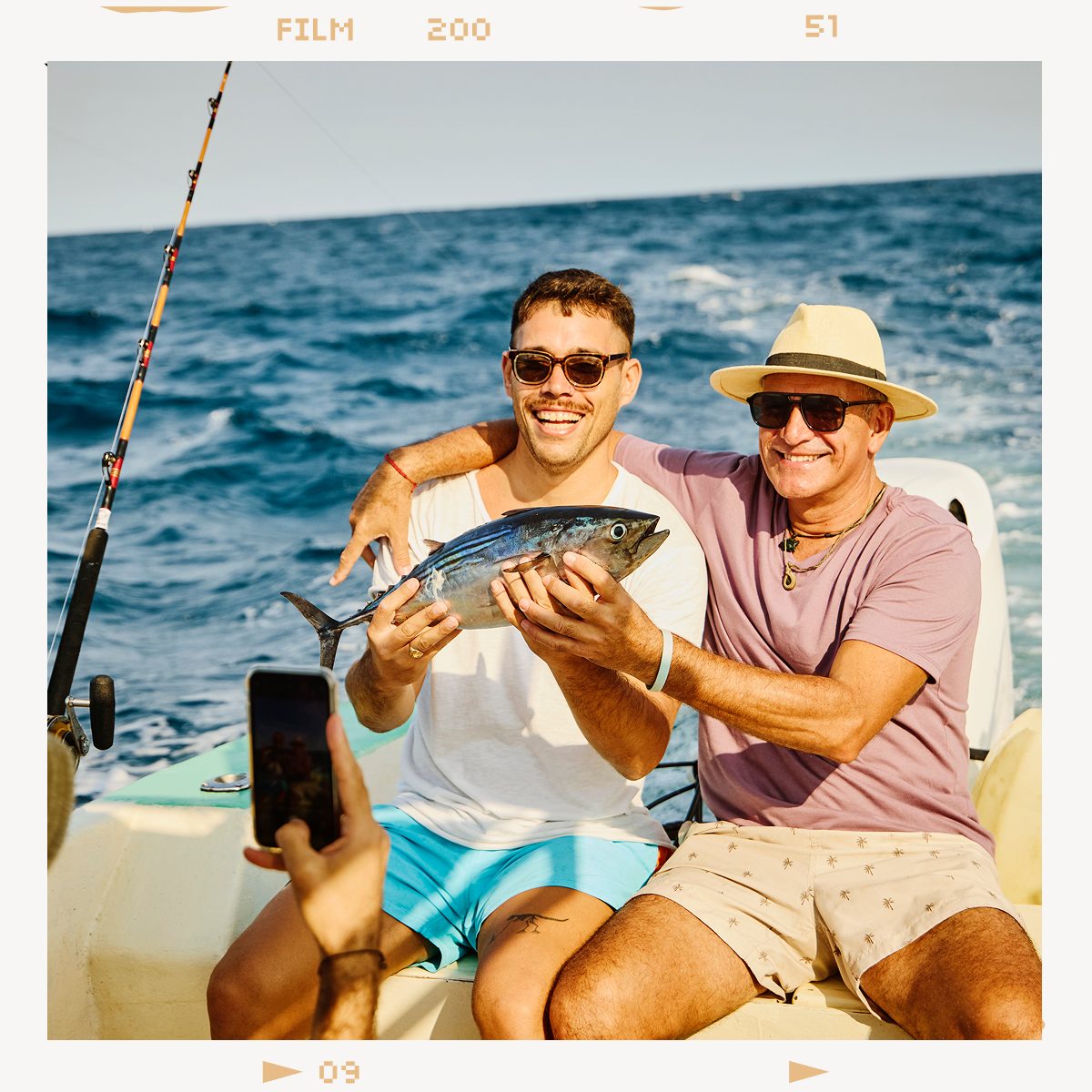 Two happy men on a boat, one holding a freshly caught fish, while another person takes a photo.
