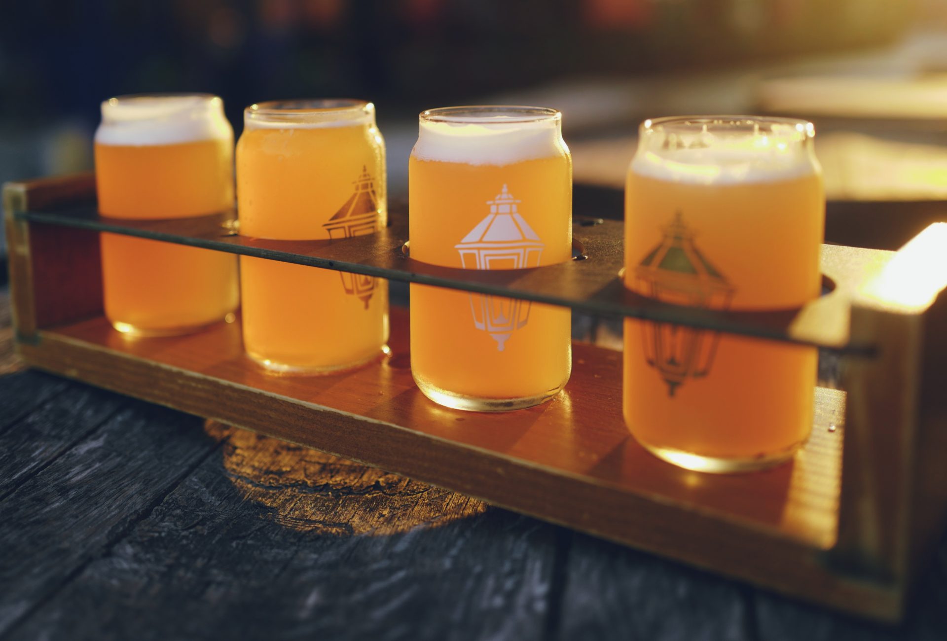 Four golden beers with foam and lantern logos in a wooden flight tray on a table.