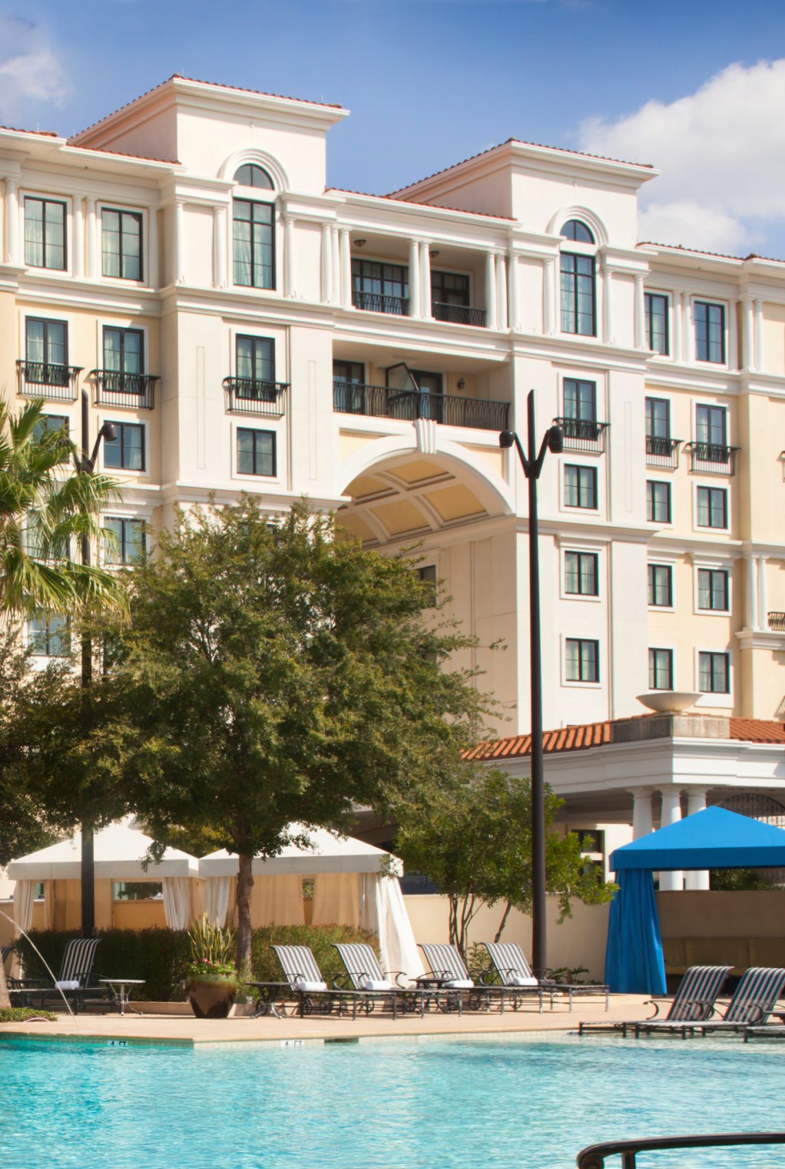 A luxurious resort hotel building with a large swimming pool, lounge chairs, and cabanas under a blue sky.