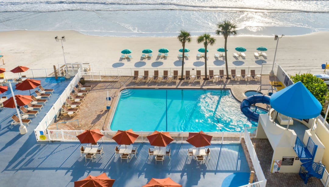 Resort pool area with umbrellas, lounge chairs, and a water slide, next to an ocean beach.