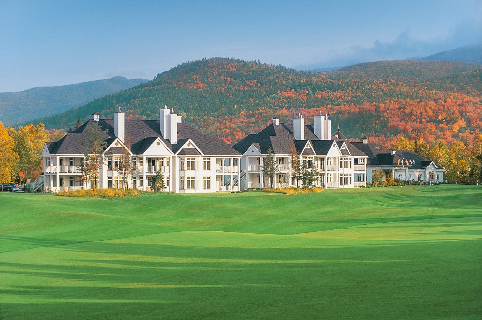 White resort buildings by a green golf course with autumn-colored mountains.
