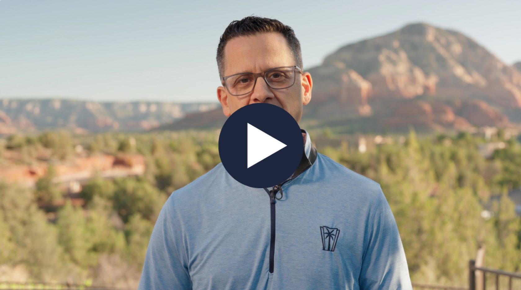 Man in glasses and blue shirt, with a play button overlay, against a natural landscape.