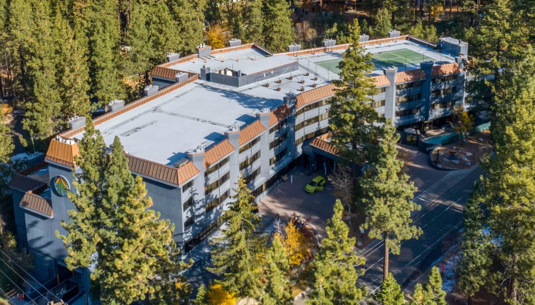Aerial view: large hotel with a rooftop tennis court, surrounded by a dense pine forest.
