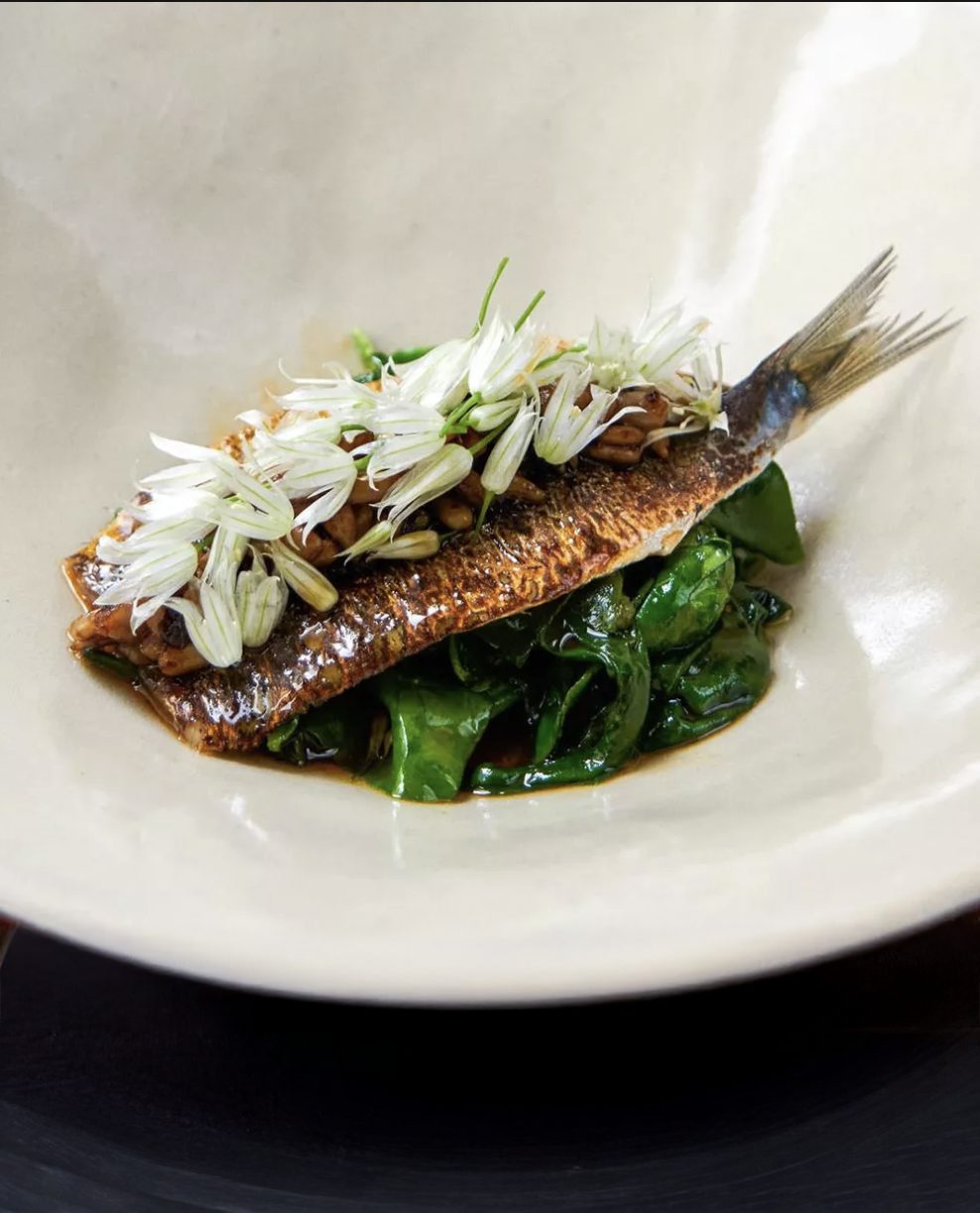 A whole crispy-skinned fish with tail, topped with white flowers & grains, on greens in a light bowl.