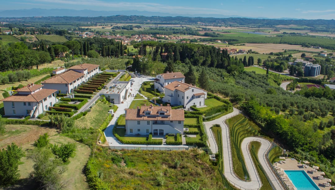 Aerial view of a resort with villas, a pool, and a winding path amidst green hills and vineyards.