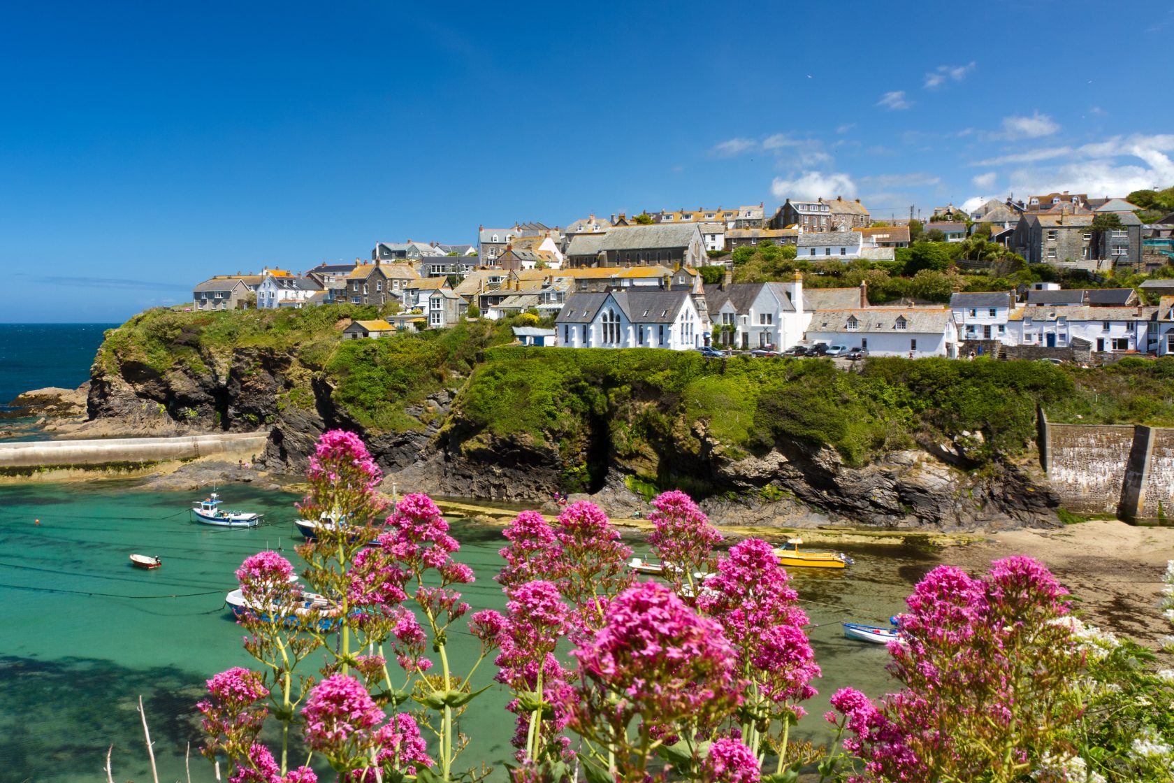 Picturesque village on cliffs overlooking a turquoise bay with boats, framed by vibrant pink flowers.