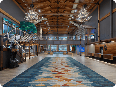 Rustic lodge lobby with large patterned rug, wood beams, bear statue, and antler chandeliers.