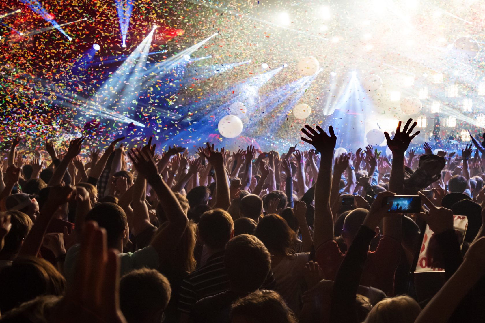 A vibrant concert scene with a crowd cheering, hands raised, under falling confetti and bright stage lights.