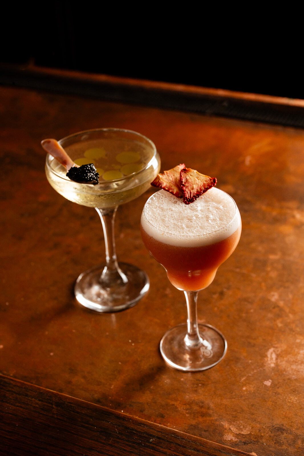 Two elegant cocktails, one with caviar and the other with foam and dried fruit, on a textured bar top.
