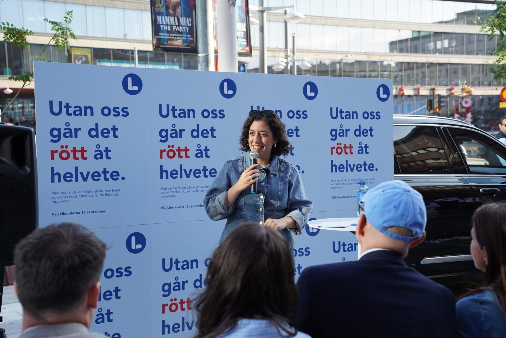 A woman speaks into a microphone at a political rally, standing in front of banners with text "Utan oss går det rött åt helvete."