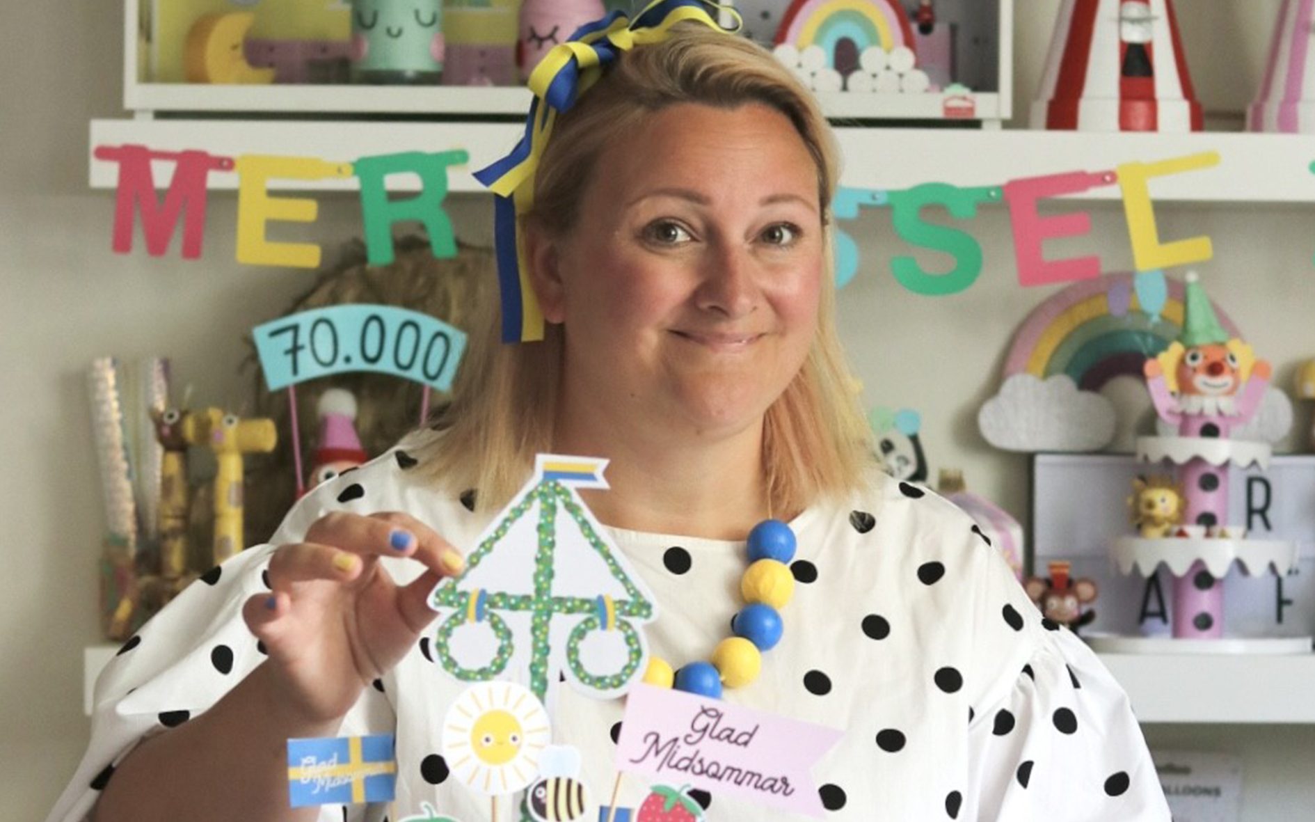 A smiling woman in a polka dot top holds a paper Midsommar pole, surrounded by colorful party decorations.
