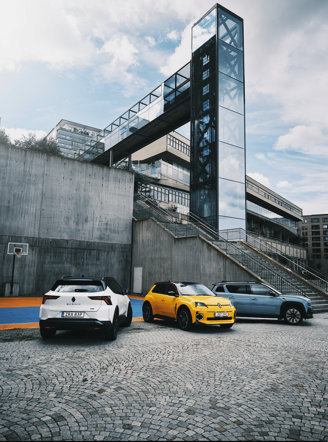 Three Renault cars (white, yellow, blue) parked on cobblestones in front of a modern building with a glass elevator.