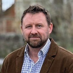 Man with beard, plaid shirt, and brown jacket looking at the camera.