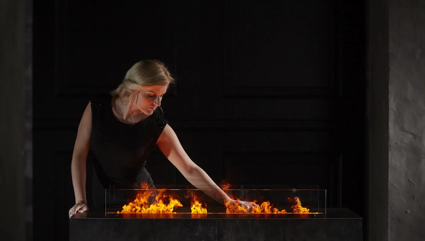 Woman in black reaching into a modern fireplace with vibrant flames.