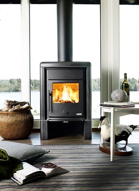 Burning fireplace by large windows with lake view, wood basket, books, and a duck statue.