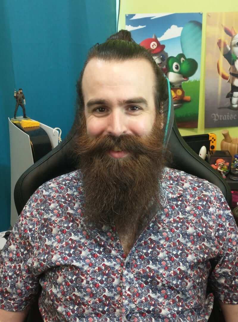 Smiling man with a large beard and hair bun, in a gaming chair, with game posters and consoles.