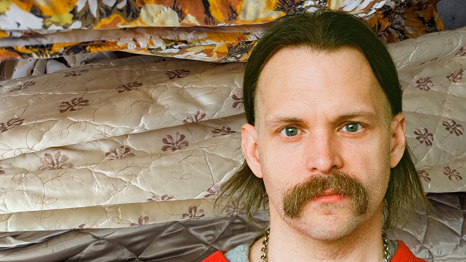 Man with a mustache and long hair against stacked, patterned blankets.