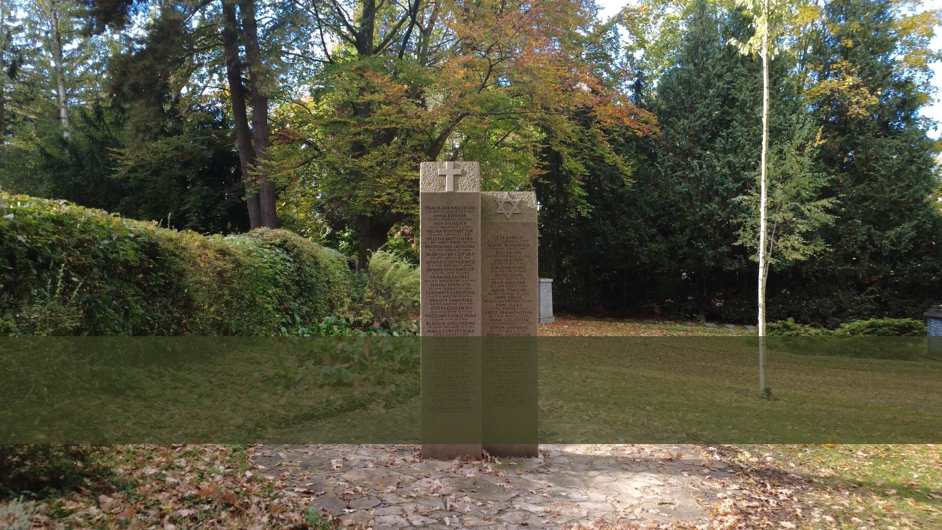 Stone memorial with a cross and Star of David, inscribed with names, surrounded by trees and grass.