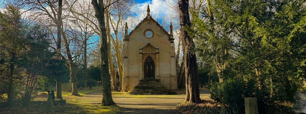 An old, ornate chapel with steps leading to its entrance, surrounded by tall trees under a partly cloudy sky.