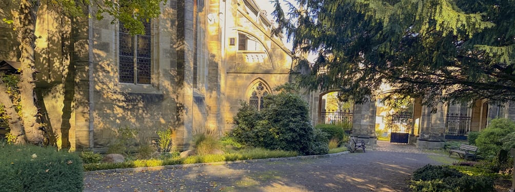 A historic stone church building and its green grounds, with dappled sunlight and shadows from large trees.