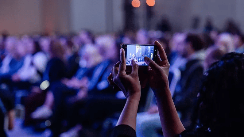 Person holding a smartphone to record an event with a blurred audience in the background.