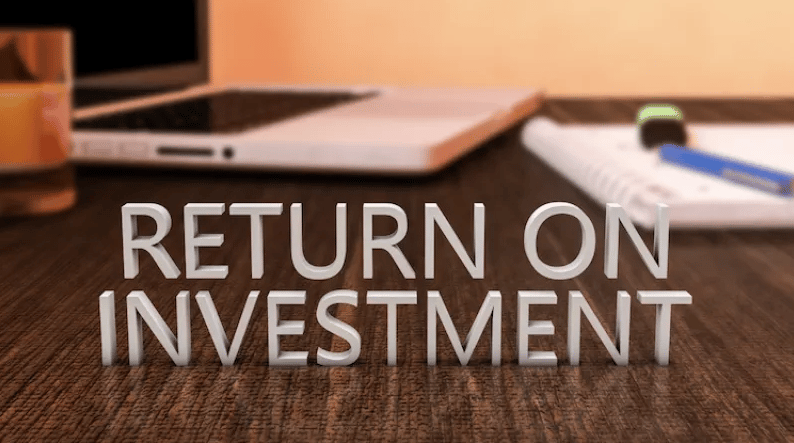 3D text 'RETURN ON INVESTMENT' on a desk with a blurred laptop and office supplies.