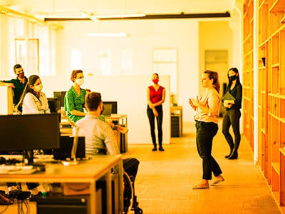 Office meeting with people, some masked. A woman stands speaking to colleagues.