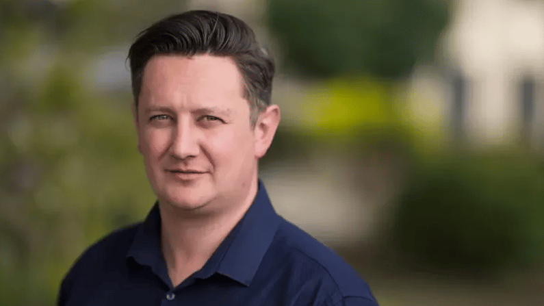 A man in a navy blue shirt looks directly at the camera with a subtle smile, blurred greenery in the background.