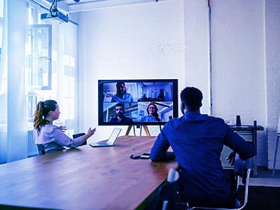 Two colleagues in an office meeting room participate in a video conference with remote team members.