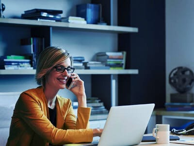 Smiling woman on phone, using laptop at desk in office.