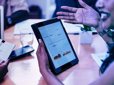 A person holds a tablet displaying data and charts during a meeting, with another person's hand gesturing.