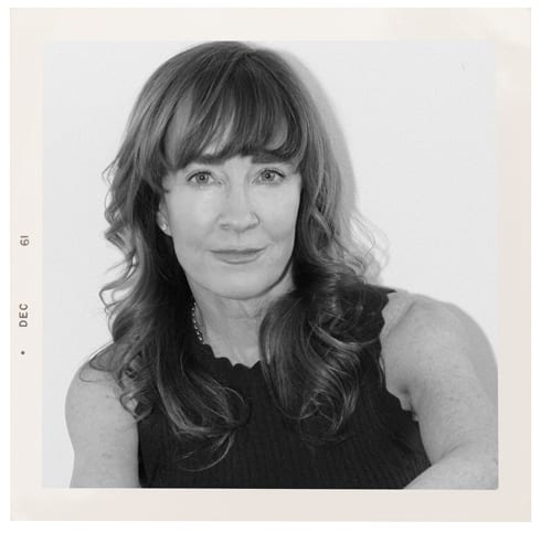 A black and white portrait of a smiling woman with long, wavy hair and bangs, wearing a dark top.