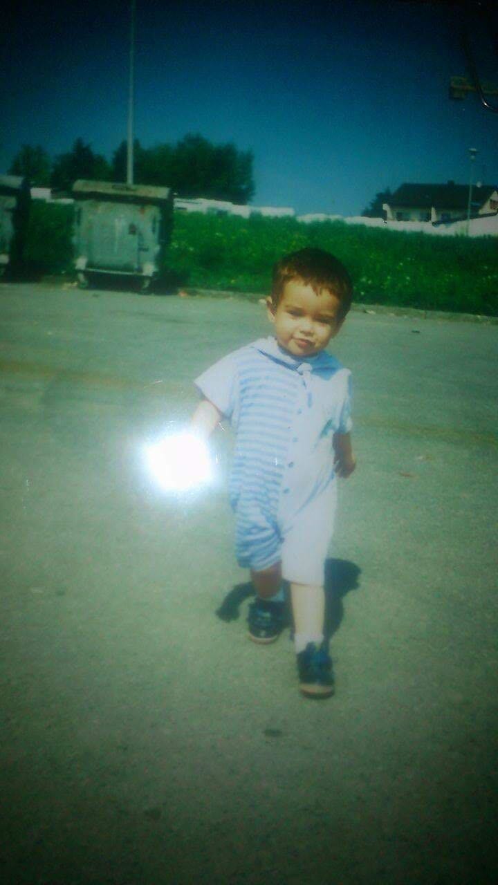 A smiling toddler in a blue and white striped romper walks on pavement, with green hills and a bright light flare.