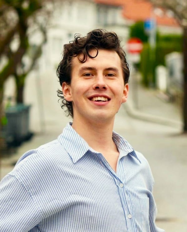 Smiling young man with dark curly hair in a light blue striped shirt.