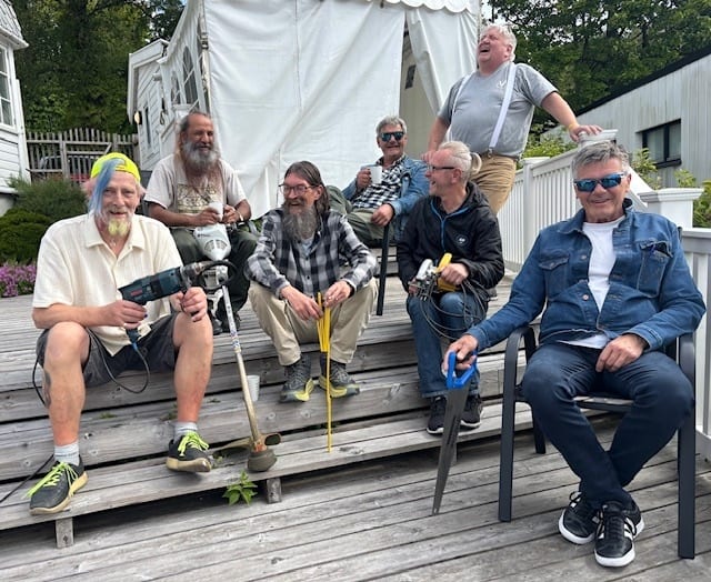 Seven men, some with tools, sitting on a deck, laughing and smiling.