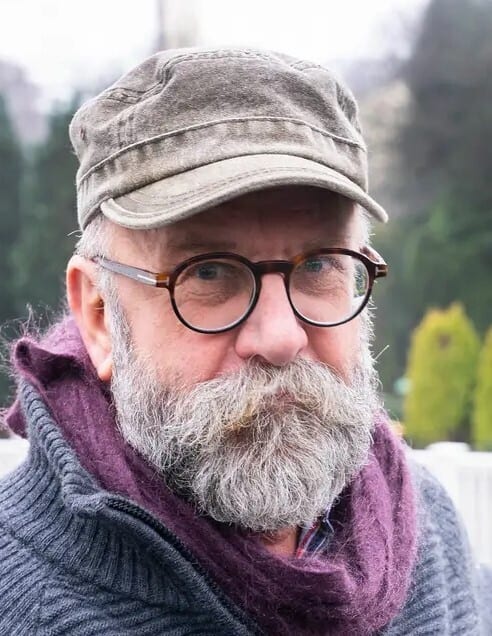 Close-up portrait of an older man with glasses, a grey beard, cap, and purple scarf.
