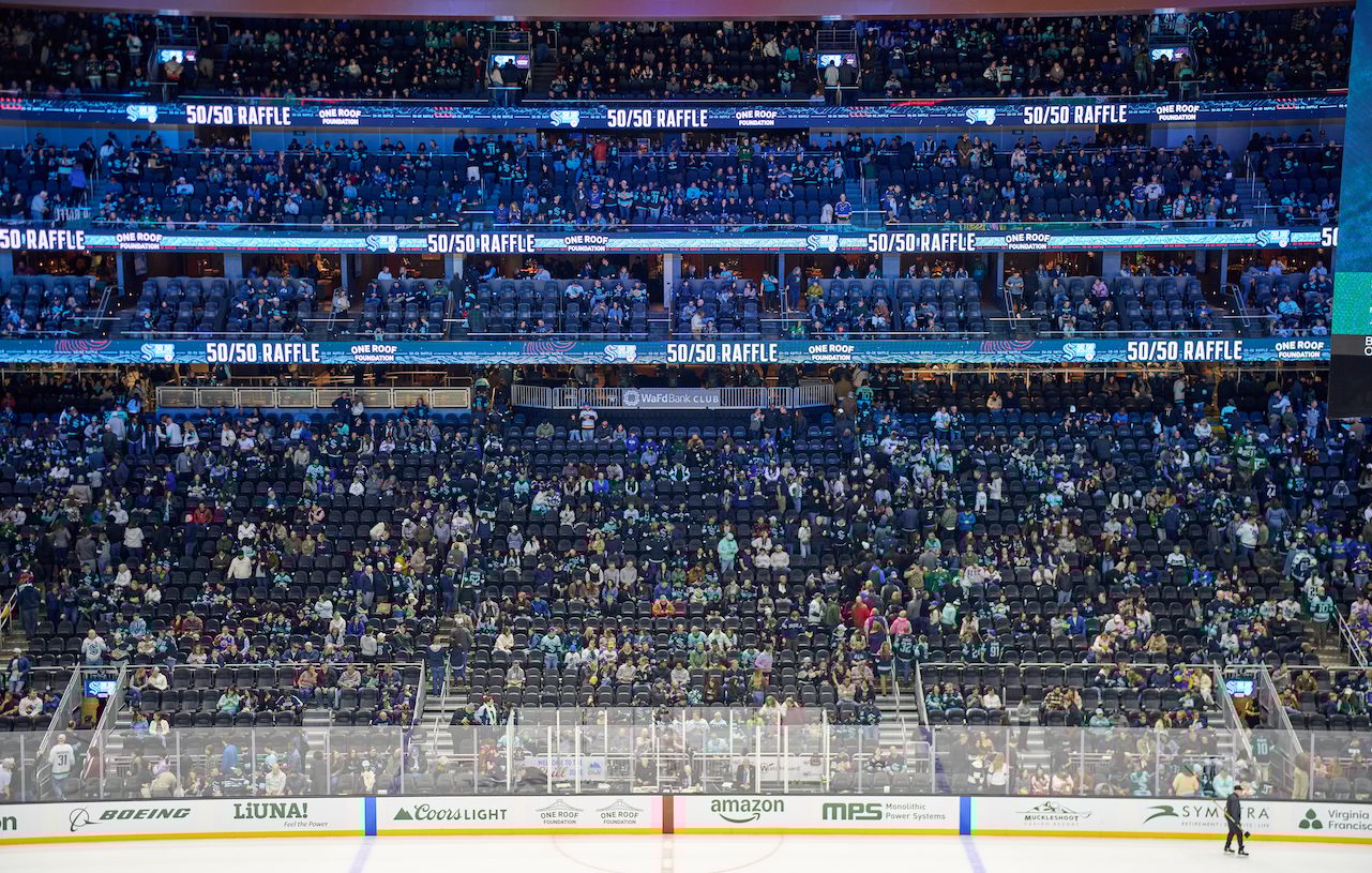Hockey arena interior with spectators, digital 50/50 Raffle signs, and rinkside sponsor ads.