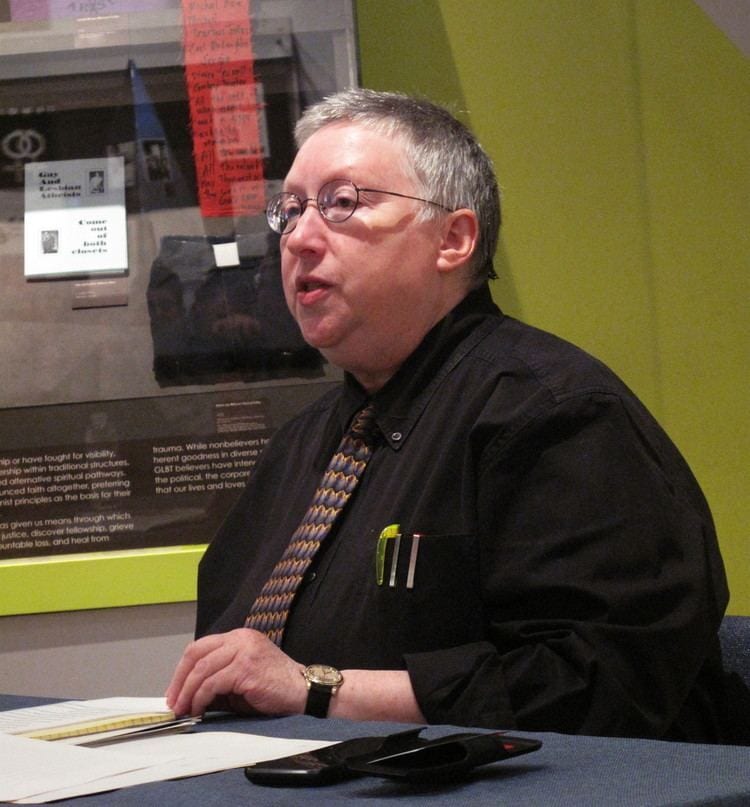 A person with short grey hair and glasses, wearing a black shirt and tie, speaks while seated.