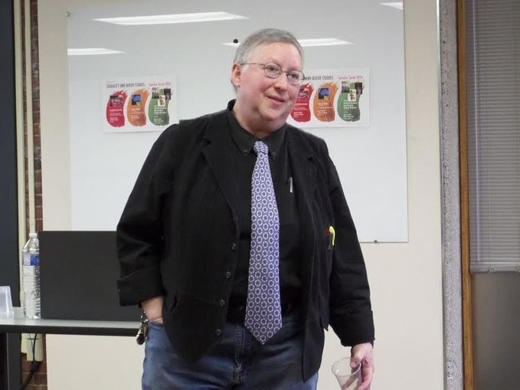 Smiling person in glasses, black blazer, and blue patterned tie, standing.