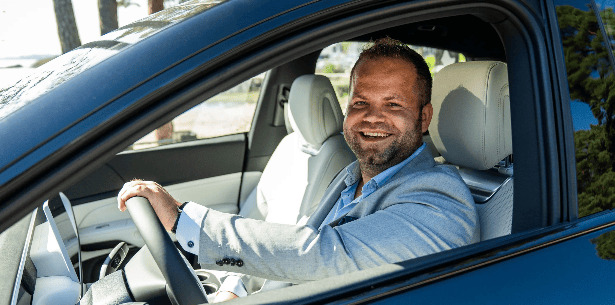 Man smiling in a car with hands on the steering wheel.