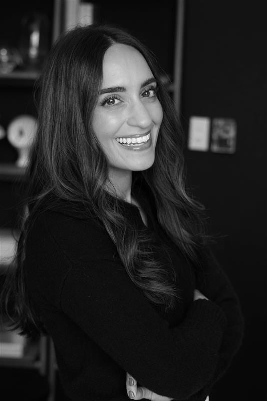 Smiling woman with long dark hair and a black top, arms crossed, in black and white.