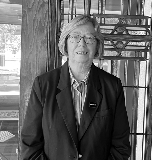 Black and white portrait of a smiling woman in glasses and a blazer, standing before a decorative window.